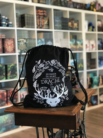 Das Produktbild zeigt einen schwarzen Turnbeutel.  Das Drachenmond-Logo (ein fliegender Drache vor einem Vollmond) ist mittig auf den Beutel gedruckt. Unter dem Logo steht "Sei immer du selbst, außer du kannst ein Drache sein". Unten am Beutel ist ein großer weißer Drache mit Hörnern abgebildet. Von diesen Hörnern aus ziehen sich verschiedene weiße Blätter und Ranken um das Logo und die Schrift herum.